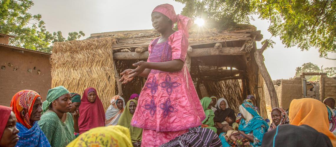 A woman standing in the middle of a group of women gathered sitting on the ground in a settlement